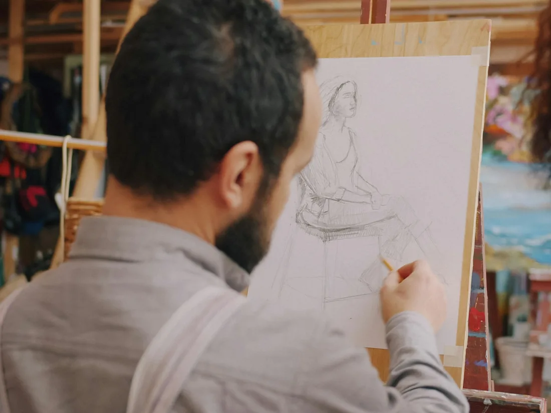 Persona dibujando a lápiz una figura sentada sobre un papel sujeto a un caballete de madera en un taller